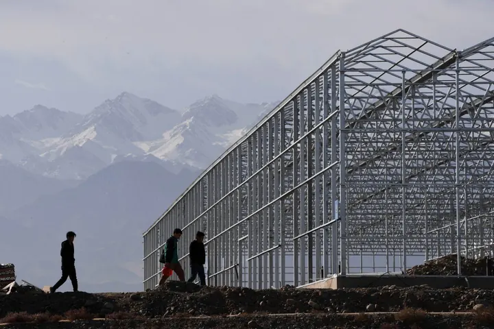 Figures walking past a long greenhouse structure beneath a mountain range