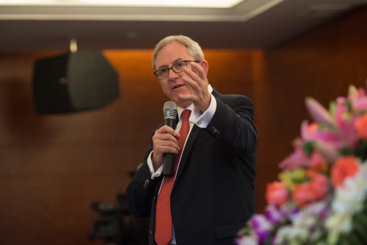 David Thomas speaking into a handheld microphone at a banquet-hall event, flowers on the table in the foreground