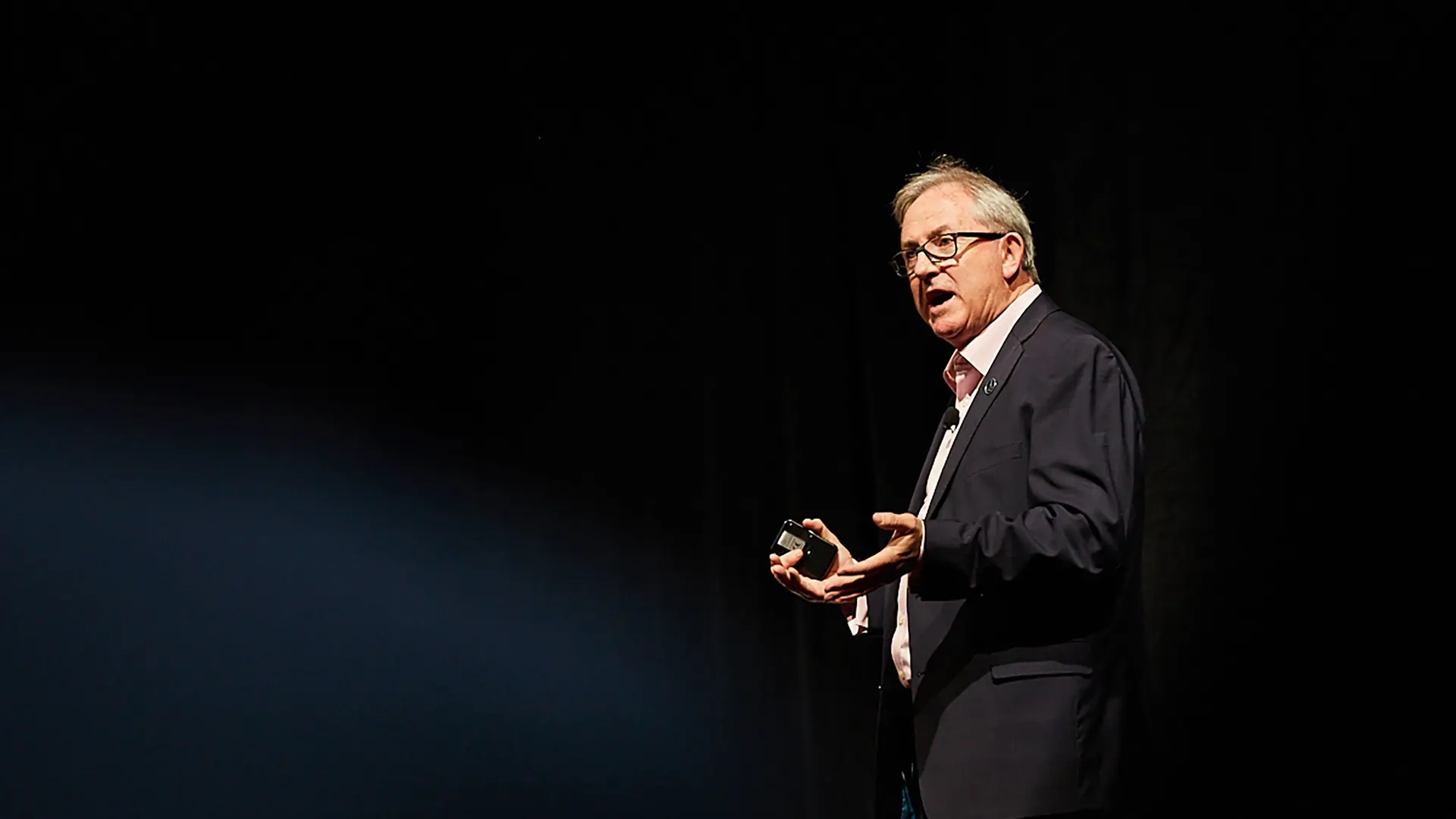 David Thomas speaking on a darkened conference stage, clicker in hand