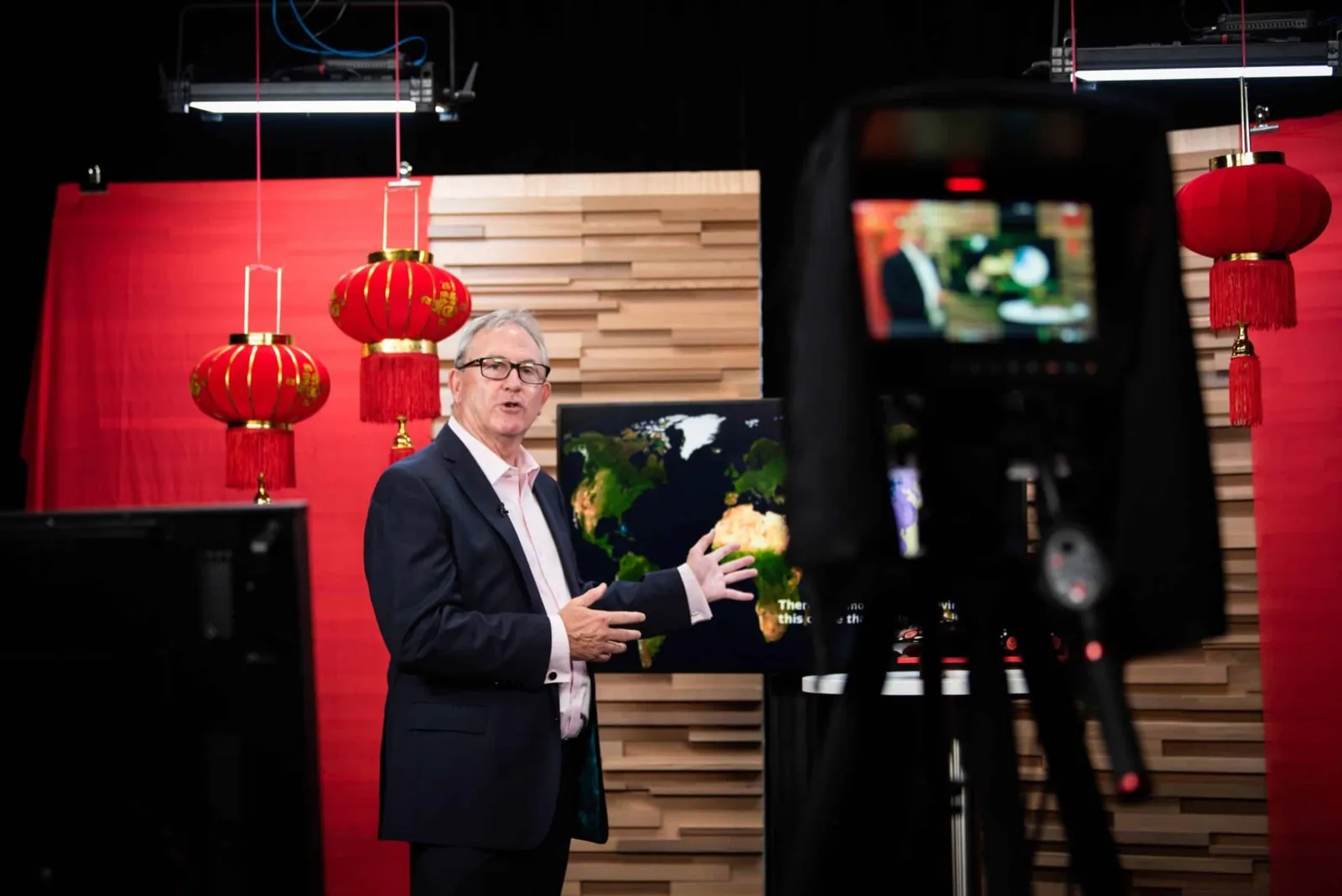 David Thomas presenting to camera on a broadcast set, red lanterns and a world map behind him